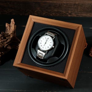 May include: A wooden watch winder with a silver wristwatch. The watch has a white face with black Roman numerals. The winder is square with a black mesh interior and a dark wood finish. A pinecone and driftwood are in the background.