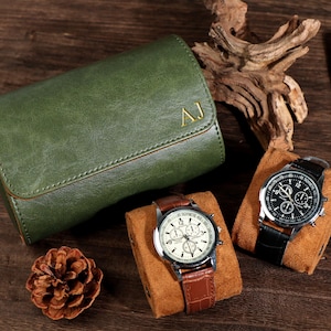 Könnte beinhalten: Draufsicht auf ein grünes Leder-Uhrenetui mit goldenen Initialen, zwei Uhren auf braunen Ständern und eine Zigarre. Eine Uhr hat ein braunes Lederarmband, die andere ein schwarzes Lederarmband. Ein Tannenzapfen und Treibholz sind ebenfalls vorhanden.