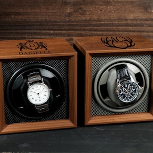 May include: Two wooden watch display cases with watches inside. One case is engraved with "DANIELLE" and a crest, the other with "AG" and antlers. The watches have silver and black bands and white and black faces.