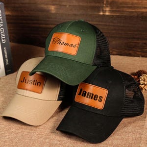 Peut inclure: Trois casquettes de camionneur personnalisées, vertes, beiges et noires. Chaque casquette comporte un écusson en cuir avec un nom : "Thomas", "Justin" et "James". Les casquettes ont des dos en filet et des visières incurvées.
