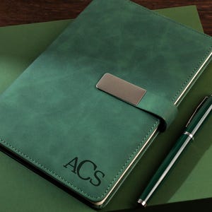 Könnte beinhalten: Ein grünes Lederjournal mit silbernem Verschluss und passendem Stift. Das Journal hat die Initialen "ACS" auf dem Einband eingeprägt. Stift und Journal liegen auf einer grünen Oberfläche.