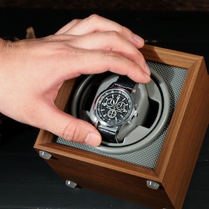 May include: A wooden watch winder with a silver and black watch. The watch has a black leather band and a black face with silver accents. The winder is square and made of dark wood.