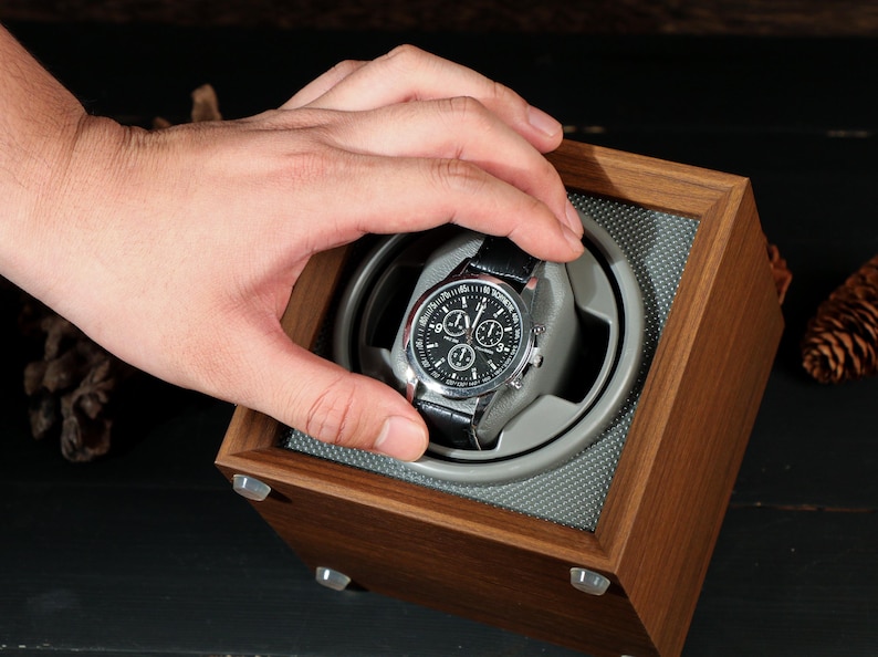 May include: A wooden watch winder with a silver and black watch. The watch has a black leather band and a black face with silver accents. The winder is square and made of dark wood.