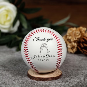 Könnte beinhalten: Ein weißer Baseball mit roten Nähten, personalisiert mit dem Text "Thank you", zwei Sektgläsern und den Namen "Julia & Chris" mit dem Datum "09.17.22". Der Baseball liegt auf einer kleinen Holzscheibe.