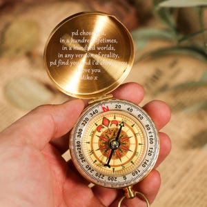 May include: A gold-coloured compass with an open lid, displaying engraved text. The compass face shows directional markings and a needle. The lid's text reads "pd chose you... I love you Miko x".