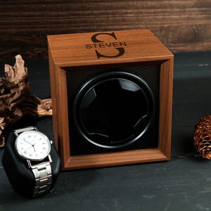 May include: A wooden watch winder with the name "STEVEN" and a large initial on the top. A silver wristwatch with a white face sits on a black stand. A pinecone and driftwood are also in the image.
