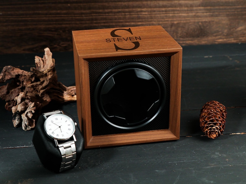 May include: A wooden watch winder with the name "STEVEN" and a large initial on the top. A silver wristwatch with a white face sits on a black stand. A pinecone and driftwood are also in the image.