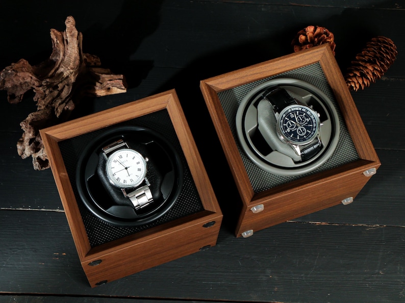 May include: Two wooden watch winders with watches inside. One watch has a white face and silver band, the other has a dark blue face and black band. The winders are square with a dark interior.