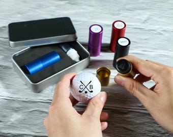 Könnte beinhalten: Ein weißer Golfball mit einem schwarzen Design aus gekreuzten Golfschlägern und den Buchstaben "C B". Der Golfball wird in einer Hand gehalten. Im Hintergrund befinden sich außerdem mehrere kleine, bunte Behälter.