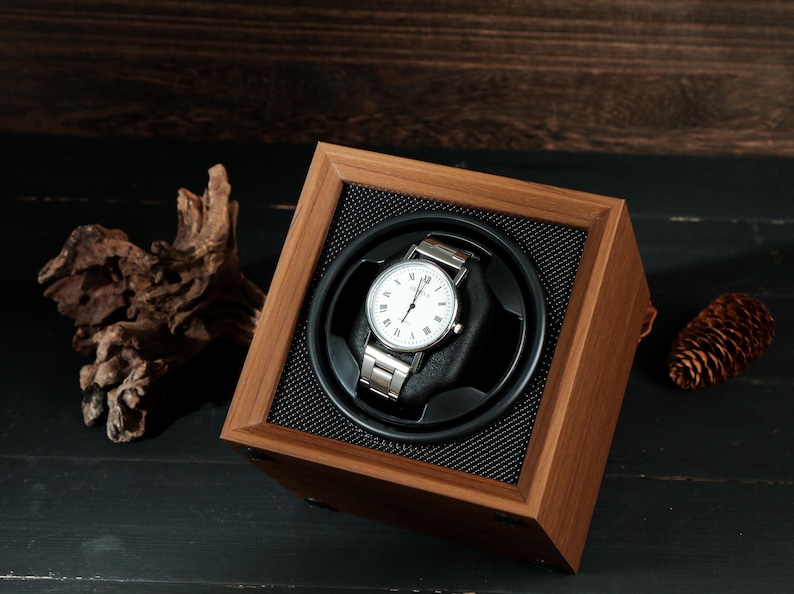 May include: A wooden watch winder with a silver wristwatch. The watch has a white face with black Roman numerals. The winder is square with a black mesh interior and a dark wood finish. A pinecone and driftwood are in the background.