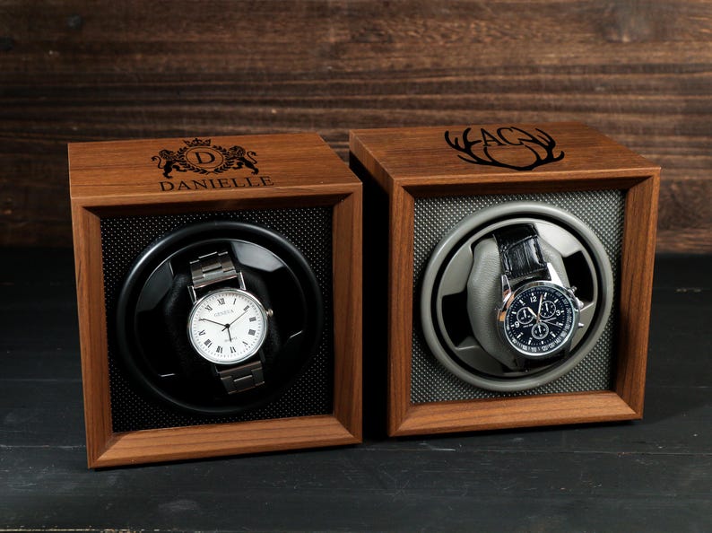 May include: Two wooden watch display cases with watches inside. One case is engraved with "DANIELLE" and a crest, the other with "AG" and antlers. The watches have silver and black bands and white and black faces.