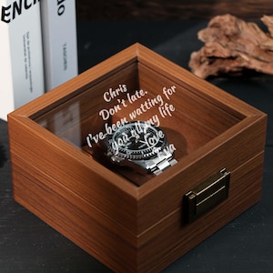 May include: A wooden watch display box with a glass top, containing a silver wristwatch with a black face. The box has a brass clasp and an inscription that reads "Chris, Don't late. I've been waiting for you all my life, Love".