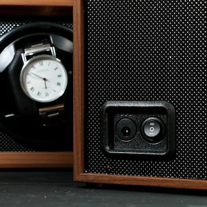 May include: A wooden watch winder with a black interior and a silver wristwatch with a white face. The winder has a perforated black front panel and a power button.