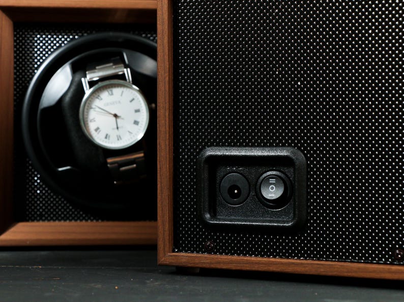 May include: A wooden watch winder with a black interior and a silver wristwatch with a white face. The winder has a perforated black front panel and a power button.