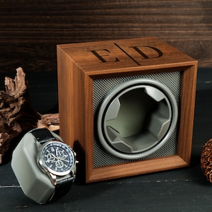 May include: A wooden watch winder with a watch on a gray stand. The winder has a square frame with a gray interior and a circular opening. The initials "E D" are engraved on the top. A black leather watch with a silver face is on the stand.