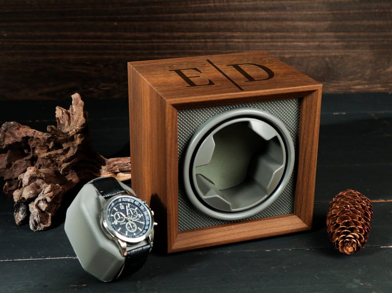 May include: A wooden watch winder with a watch on a gray stand. The winder has a square frame with a gray interior and a circular opening. The initials "E D" are engraved on the top. A black leather watch with a silver face is on the stand.