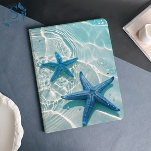 Può includere: Un quaderno blu e bianco a tema spiaggia. La copertina presenta due stelle marine blu su uno sfondo azzurro con increspature d'acqua e bolle. Il quaderno ha un design pulito e moderno.