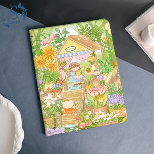 May include: A rectangular notebook featuring a colorful illustration of a small wooden structure surrounded by sunflowers, tulips, and various green plants. A cartoon character stands inside the structure, while a small bear figure sits on the steps.