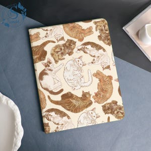 May include: A rectangular notebook with a cream-colored cover featuring a repeating pattern of various cat illustrations in shades of brown and white. The cats are depicted in playful poses.