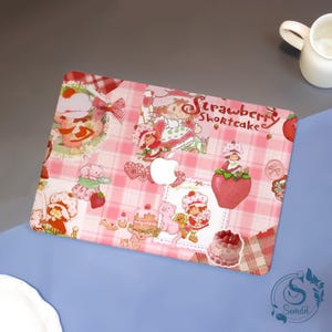 May include: A pink and white plaid laptop cover featuring Strawberry Shortcake characters and the text "Strawberry Shortcake". The cover has various illustrations of the character, strawberries, and desserts. The Apple logo is visible.