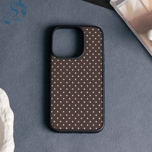 May include: A black phone case featuring a brown polka dot design. The case has a black edge and a camera aperture. The small, light pink polka dots are evenly spaced. The phone case is on a grey surface.