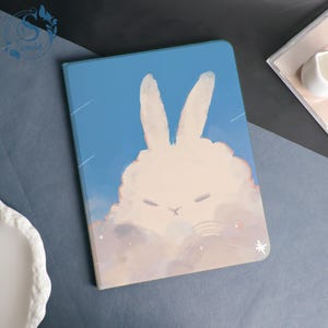 May include: A blue and white tablet case featuring a whimsical illustration of a sleeping bunny in a cloud. The design includes a light blue sky background with shooting stars and a small snowflake.
