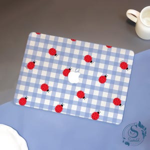 Peut inclure: Une coque d'ordinateur portable avec un motif vichy bleu clair et blanc, ornée d'illustrations de coccinelles rouges. Le boîtier présente un logo Apple blanc au centre.