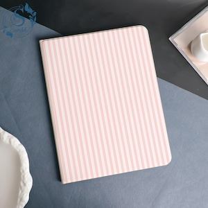 May include: A rectangular notebook with a pink and white striped pattern. The notebook is closed and has rounded corners. The cover is a light pink colour with vertical white stripes. The notebook is on a dark grey surface.