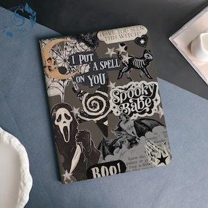 Può includere: Un taccuino grigio con un collage a tema Halloween. La copertina presenta un teschio, un gatto scheletro e le scritte "I PUT A SPELL ON YOU" e "Spooky Babe".
