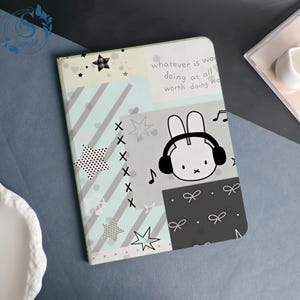 May include: A rectangular notebook with a multi-panel design featuring light blue diagonal stripes, a grey section with a white rabbit wearing black headphones, and a dark grey panel with white bow patterns. Text reads: whatever is worth doing at all is worth doing well.
