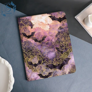 Pode incluir: Um caderno retangular com um design temático de Halloween. A capa apresenta um céu roxo e rosa com lua cheia, silhuetas de morcegos e árvores douradas. O caderno está sobre uma superfície cinza.