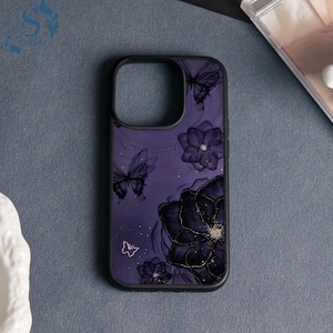 May include: A phone case with a black border and a purple background. The case features a floral and butterfly design in black and gold. The flowers have gold glitter accents, and the butterflies are outlined in black.
