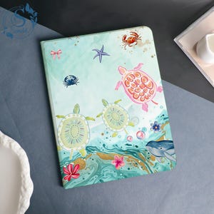 May include: A rectangular tablet case featuring a light green and blue watercolor background with illustrations of two sea turtles, a pink sea turtle, a blue whale, a red crab, a small blue crab, a purple starfish, pink flowers, and a small pink bow.