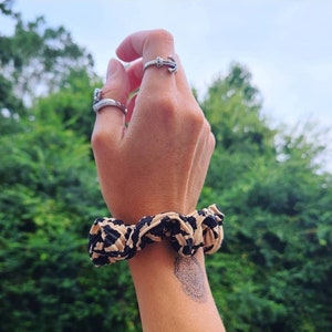 May include: A black and beige scrunchie with a textured fabric. The scrunchie is on a person's wrist, and there is a small tattoo on the wrist.