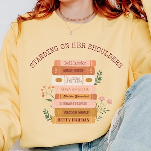 May include: A yellow sweatshirt with the text "STANDING ON HER SHOULDERS" and a stack of books with names like Audre Lorde and Maya Angelou. The sweatshirt has a crew neck and long sleeves. The word "BUTTER" is in a circle on the bottom left.
