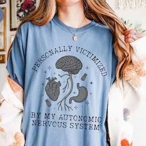 May include: A pale blue t-shirt featuring the words "PERSONALLY VICTIMIZED BY MY AUTONOMIC NERVOUS SYSTEM" and a graphic of a brain, heart, and stomach. The shirt has a round neck and is a solid colour.
