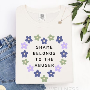 Könnte beinhalten: Elfenbeinfarbenes Comfort Colors T-Shirt mit dem Text "SHAME BELONGS TO THE ABUSER" in einem floralen Herzdesign. Das gefaltete Shirt liegt neben einer blauen Jeans und einer goldenen Halskette.