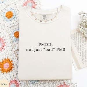 Può includere: Felpa color avorio con la scritta "PMDD: not just 'bad' PMS" in nero. Una delicata collana e un libro con fiori sono sulla destra. La felpa è su una superficie bianca con una colorata coperta all'uncinetto.