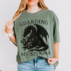 May include: Moss green t-shirt with a black dragon graphic guarding spoons. The text "GUARDING MY SPOONS" is printed above and below the dragon. The shirt is a short-sleeved, crew neck style.