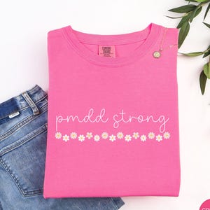 May include: A folded, bright pink t-shirt with the words "pmdd strong" in a cursive font, and a row of small daisy graphics. A gold necklace with a pendant rests on the shirt. A pair of blue jeans is partially visible.