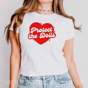 Puede incluir: Camiseta blanca con un gráfico de corazón rojo. El corazón contiene el texto "Protect the Dolls" en una fuente retro. La camiseta se combina con vaqueros azules.
