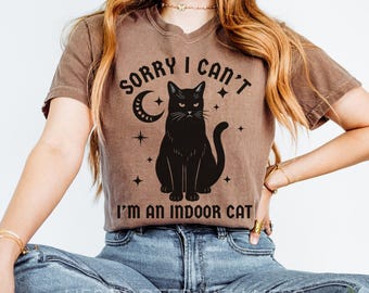 Könnte beinhalten: Espressofarbenes T-Shirt mit dem Text "SORRY I CAN'T I'M AN INDOOR CAT" und einer schwarzen Katzen-Grafik. Das Design beinhaltet einen Mond und Sterne. Das Shirt wird mit einer blauen Jeans getragen.