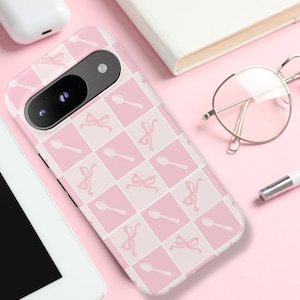 Op de afbeelding: Een roze telefoonhoesje met een geruit patroon van witte en roze vierkanten. Elk vierkant heeft een roze lepel en een roze strik.