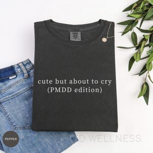 Könnte beinhalten: Ein dunkelgraues T-Shirt mit dem weißen Text "cute but about to cry (PMDD edition)". Das Shirt ist gefaltet und auf einer weißen Oberfläche platziert, mit einer Jeans und einer Halskette. Grüne Blätter sind im Hintergrund.