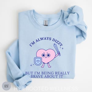 May include: Light blue sweatshirt with the text "I'M ALWAYS DIZZY" above a pink heart character holding a sword and shield. Below the heart is the text "BUT I'M BEING REALLY BRAVE ABOUT IT". The sweatshirt has a crew neck and long sleeves.