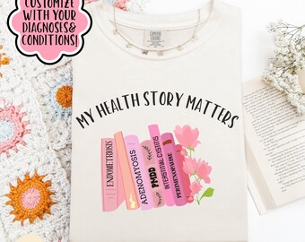 May include: Ivory-colored t-shirt with the text "MY HEALTH STORY MATTERS" and a graphic of pink books labeled with medical conditions. The shirt also features the text "CUSTOMIZE WITH YOUR DIAGNOSES & CONDITIONS!"