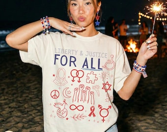 Könnte beinhalten: Beigefarbenes T-Shirt mit dem Text "LIBERTY & JUSTICE FOR ALL" in Blau und Rot, zusammen mit verschiedenen inklusiven Symbolen. Die Person trägt rot-weiß-blaue Perlenarmbänder und Ohrringe. Ein Wunderkerze ist ebenfalls im Bild zu sehen.