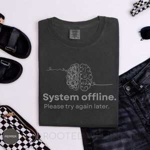 May include: Dark gray t-shirt with a white brain graphic and the text "System offline. Please try again later." The shirt is a crew neck and appears to be made of a soft, comfortable material.