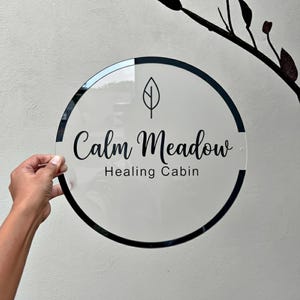 May include: A clear, round sign with a black border and the words "Calm Meadow Healing Cabin" in elegant script. A leaf graphic is above the text. The sign is held by a hand against a neutral background.