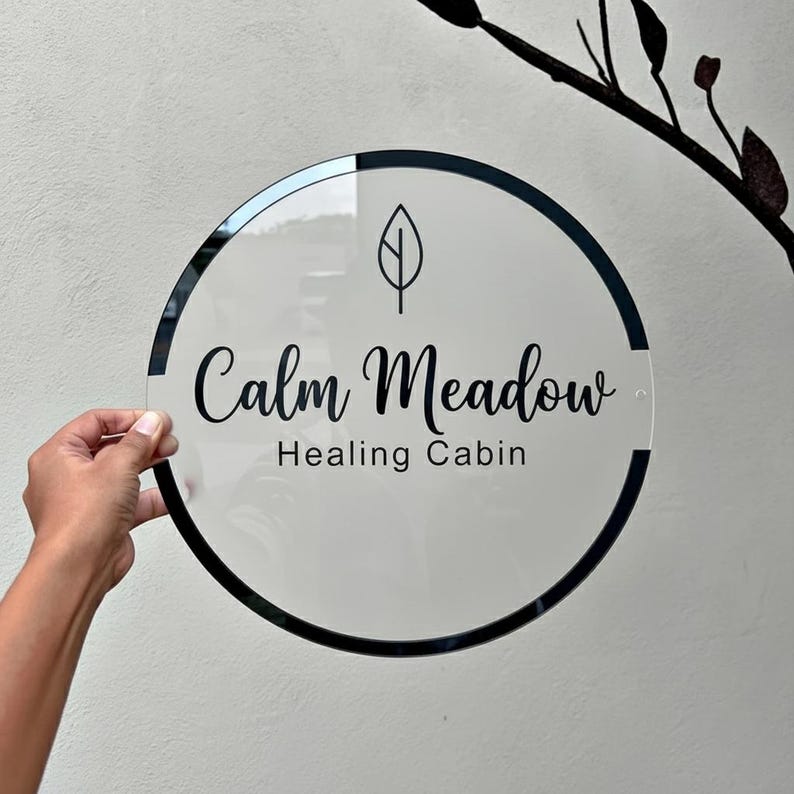 May include: A clear, round sign with a black border and the words "Calm Meadow Healing Cabin" in elegant script. A leaf graphic is above the text. The sign is held by a hand against a neutral background.